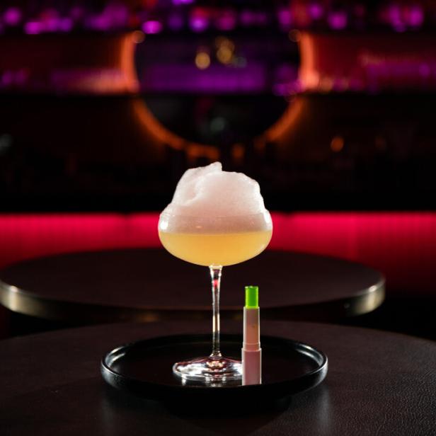 Ein Appletini-Cocktail mit Schaumhaube und einem Lippenpflegestift auf einem Tisch.