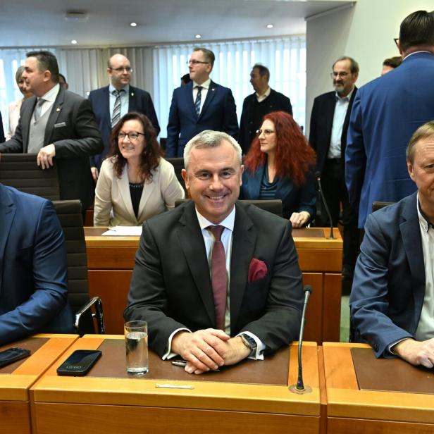 Mehrere Personen in Anzügen und Kostümen sitzen und stehen in einem Konferenzraum, einige lächeln und unterhalten sich.