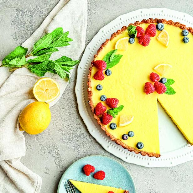 Eine Zitronentarte mit Himbeeren und Blaubeeren, ein Stück davon auf einem Teller.