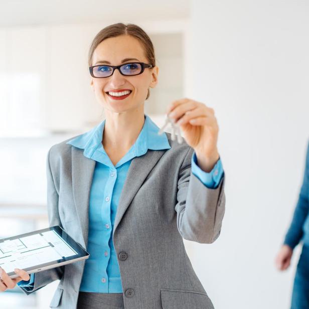 Eine Frau im Business-Blazer mit einem Schlüssel in der Hand. Im Hintergrund: Ein Paar. 