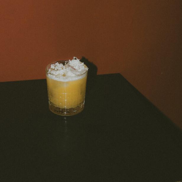 Ein gelber Cocktail mit Eiswürfeln und einer weißen Garnitur auf einem schwarzen Tisch.