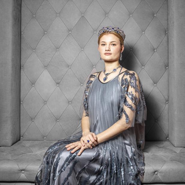 Eine junge Frau sitzt in einem grauen Kleid und Diadem auf einem Sofa.