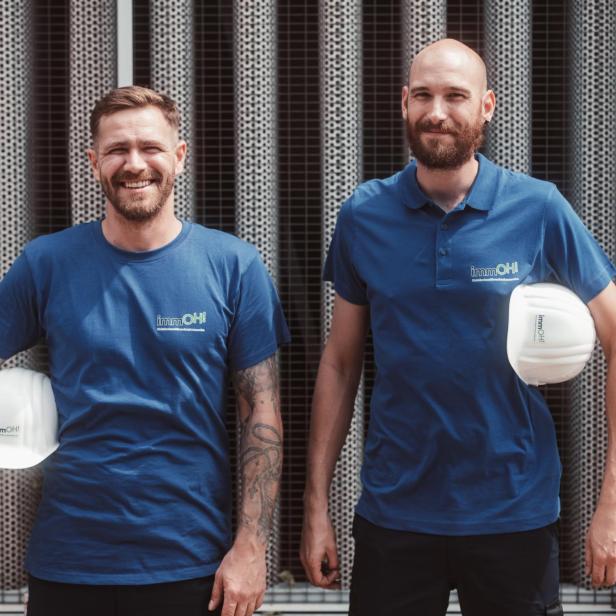 Zwei weiße Männer stehen mit blauen Shirts und weißen Helmen da. 