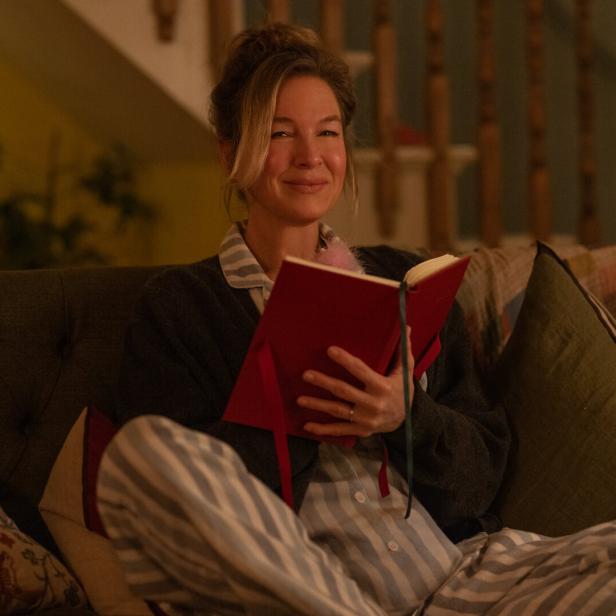 Renée Zellweger sitzt im Schlafanzug auf einem Sofa und liest in einem roten Buch.