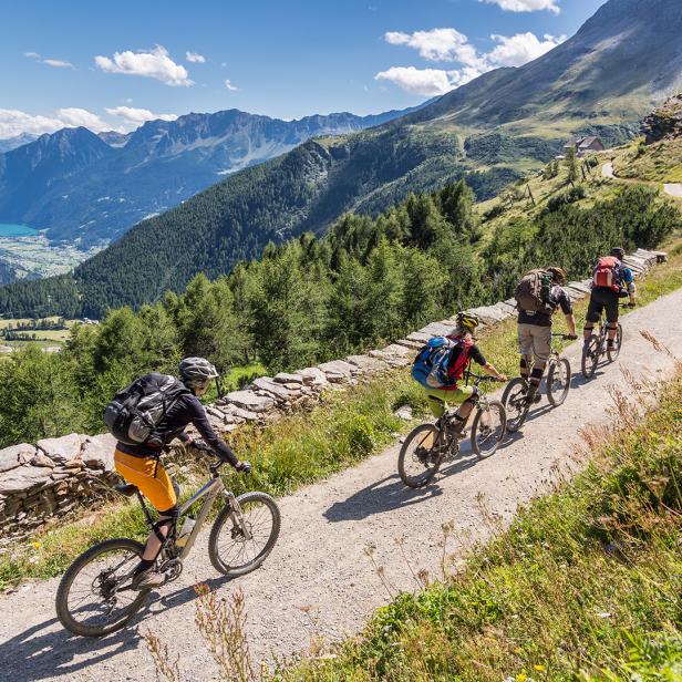 Vier Fahrradfahrer fahren einen Berg hoch. Neben ihnen ein See mit Bergen drumherum. 