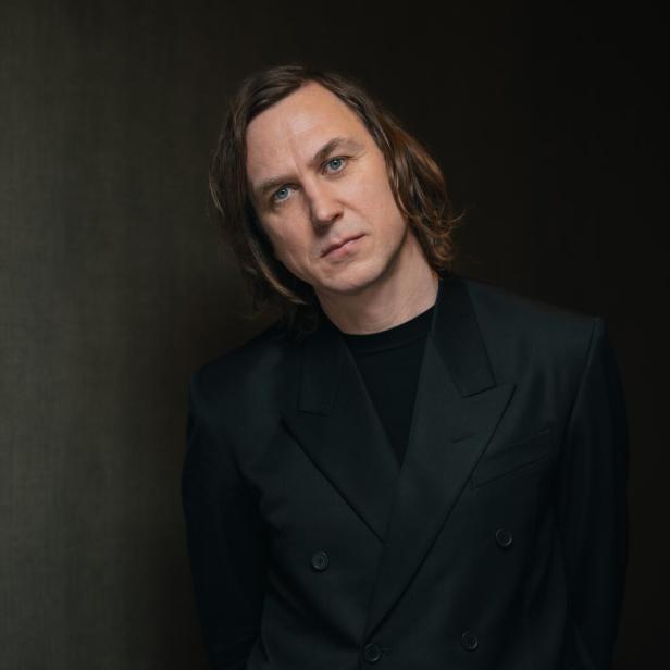 Porträt von Jarvis Cocker in einem schwarzen Anzug vor einem dunklen Hintergrund.