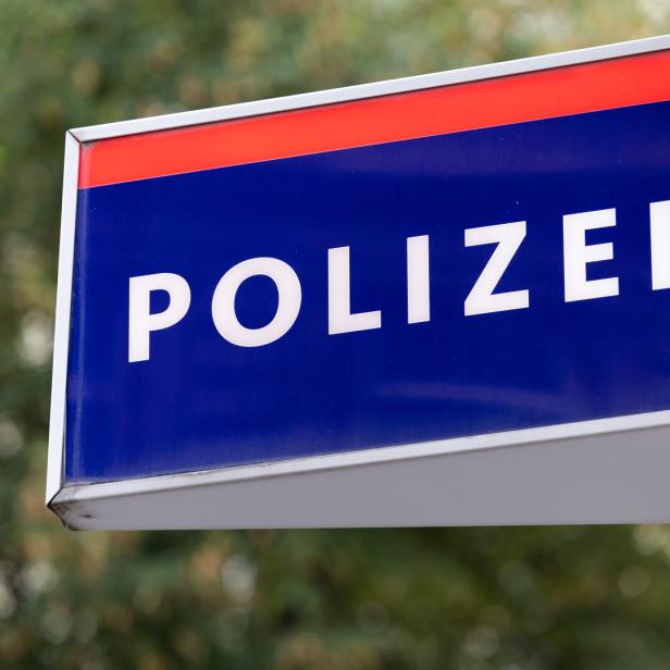 Blaues Schild mit weißer Aufschrift „Polizei“ und einem Adlerwappen, im Hintergrund unscharfe Bäume.