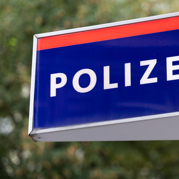 Blaues Schild mit weißer Aufschrift „Polizei“ und einem Adlerwappen, im Hintergrund unscharfe Bäume.