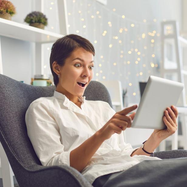 Eine Frau sitzt auf einem Sessel und schaut erstaunt auf ihr Tablet. 
