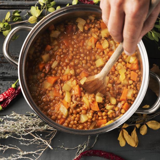 Ein Koch rührt mit einem Holzlöffel eine Linsensuppe mit Gemüse in einem Topf.