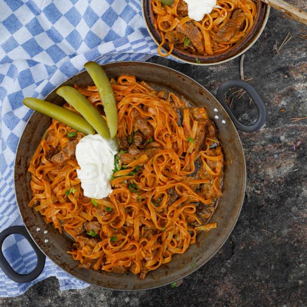 Boeuf Stroganoff mit Nudeln, saurer Sahne und Gurken in einer Pfanne und einer Schüssel.