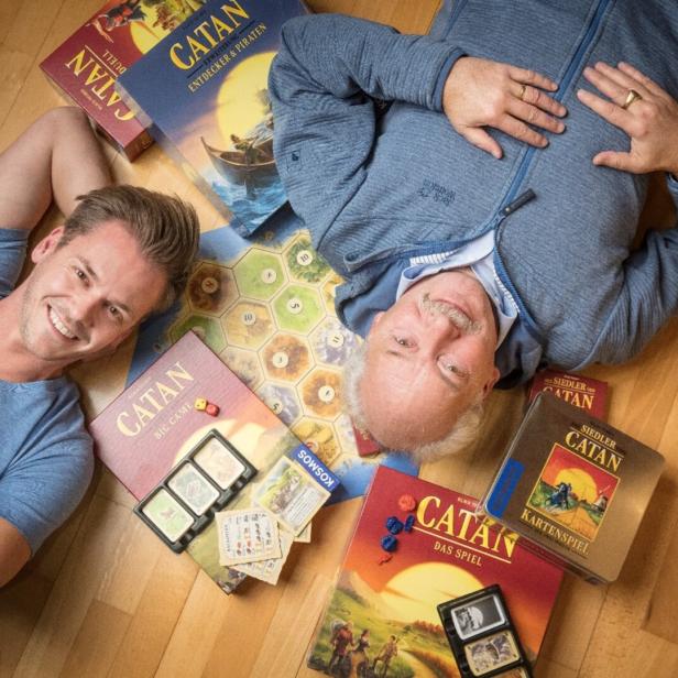 Zwei Männer liegen auf dem Boden, umgeben von verschiedenen „Catan“-Spielen.