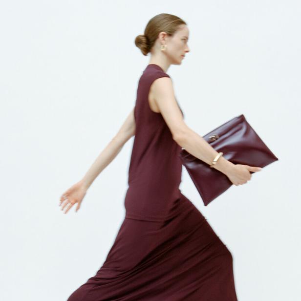 Eine Frau in einem langen, weinroten Kleid und Sandalen hält eine passende Clutch.