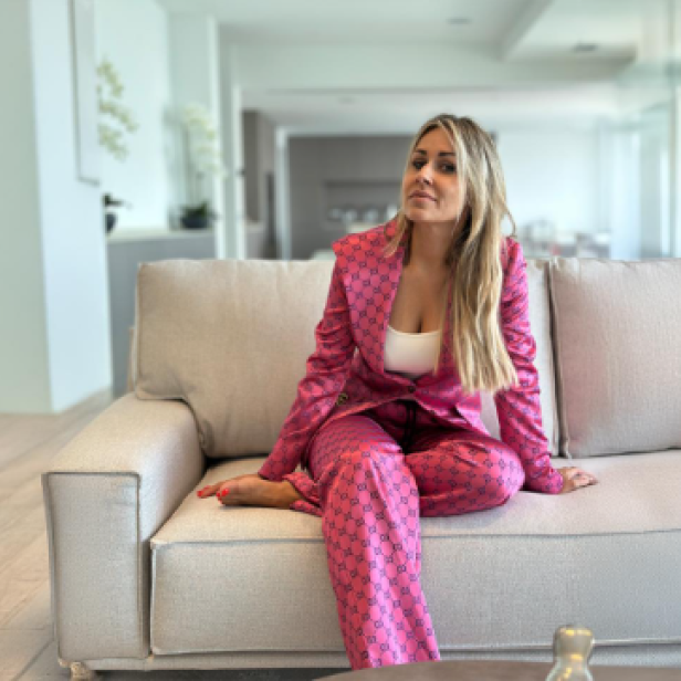 Eine blonde Frau sitzt in einem pinken Anzug auf einem beigen Sofa. 