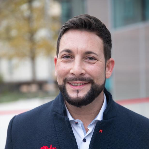 Ein lächelnder Mann mit Bart und Nelke am Revers seines Sakkos.