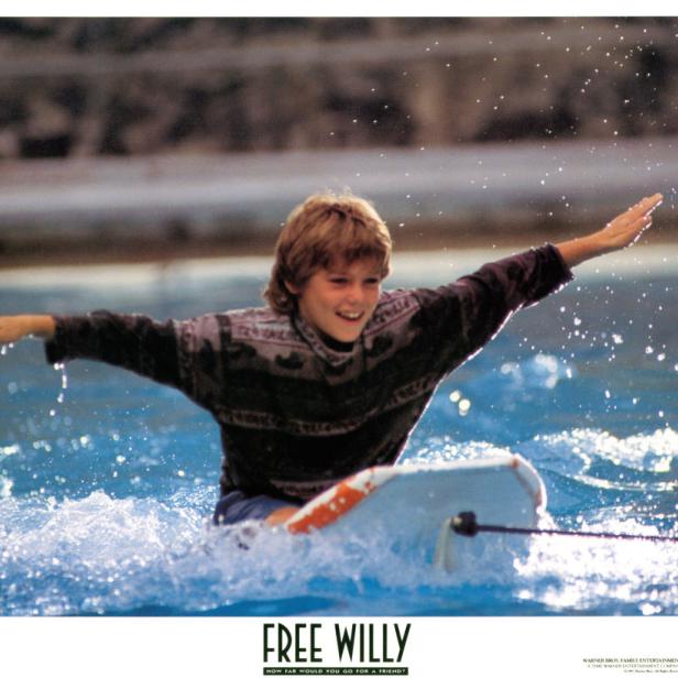 Ein lächelnder Junge reitet auf einem Surfbrett im Wasser, Szene aus dem Film „Free Willy“.