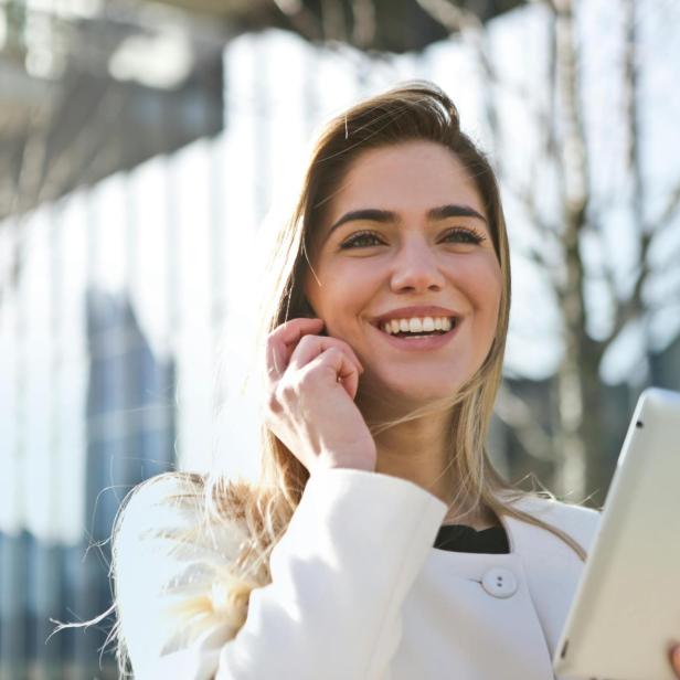 Eine blonde Frau lächelt, mit einem Handy am Ohr und einem Tablet in der anderen Hand. 
