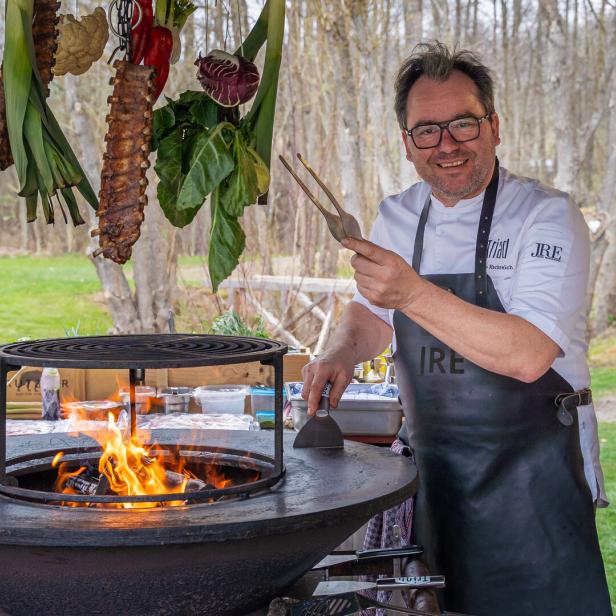 Uwe Machreich bei seinem Griller aus Platte und Feuerstelle. Darüber hängen Ripperl und Gemüse.