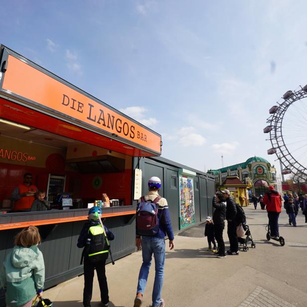 Ein Imbissstand „Die Langos Bar“ im Wiener Prater mit Menschen und dem Riesenrad im Hintergrund.
