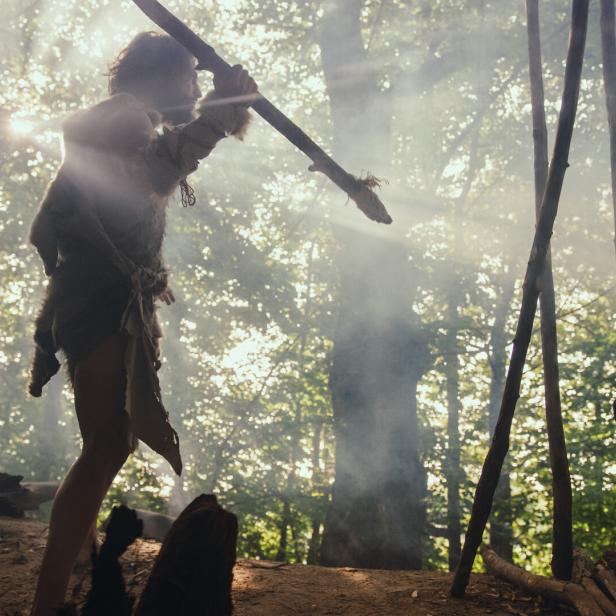 Urzeitlicher Höhlenmensch, der ein Tierfell trägt, hält einen Speer aus Stein, steht am Höhleneingang und blickt über den prähistorischen Wald, bereit.