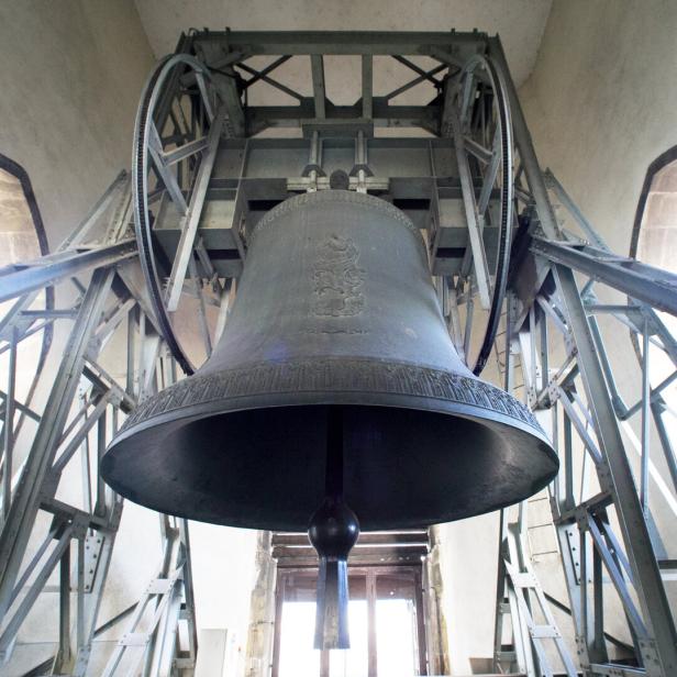 Eine große Glocke hängt in einem metallenen Glockenstuhl vor einem hellen Fenster.