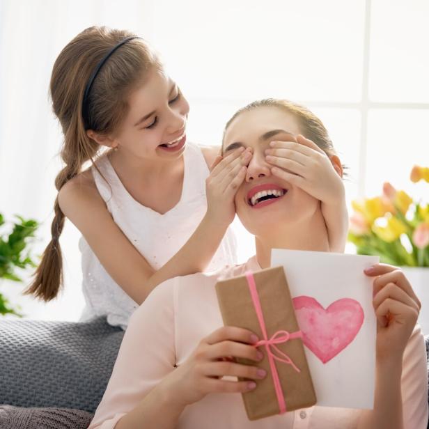 Ein Mädchen hält ihrer Mutter die Augen zu, während diese ein Geschenk und eine Karte mit einem Herz in den Händen hält.
