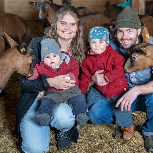Eine Familie sitzt inmitten einer Ziegenherde im Stall.