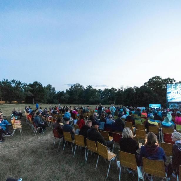 Besucher sitzen auf Stühlen im grünen vor einer Leinwand, ein Open-Air-Kino. 