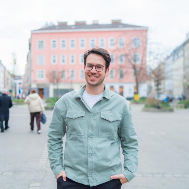Ein lächelnder Mann mit Brille steht auf einem belebten Platz in der Stadt.