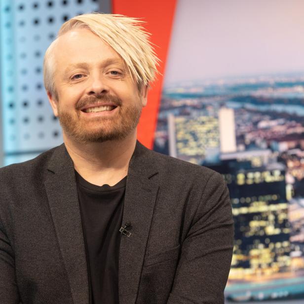 Ross Antony lächelt vor einem unscharfen Stadtbild.
