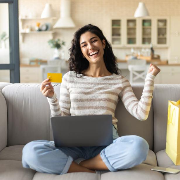 Junge Frau sitzt lachend im Schneidersitz auf einem Sofa, hält eine Kreditkarte in der einen Hand und hat einen Laptop auf dem Schoss; neben ihr stehen bunte Einkaufstaschen, im Hintergrund ist eine helle Küche zu sehen.
