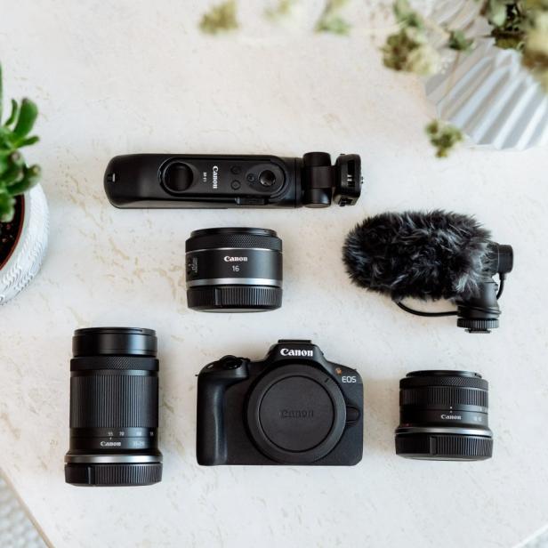 Flach gelegtes Set aus Canon-Foto- und Videozubehör auf einem hellen Tisch: in der Mitte eine Canon EOS-Kamera, umgeben von drei Objektiven, einem Mikrofon mit Windschutz, einem Handgriff mit integrierter Fernbedienung sowie dekorativen Pflanzen im oberen linken und rechten Bildbereich.
