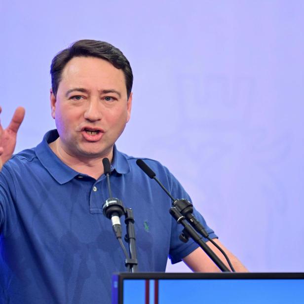 Ein Mann in einem blauen Poloshirt spricht vor Mikrofonen.