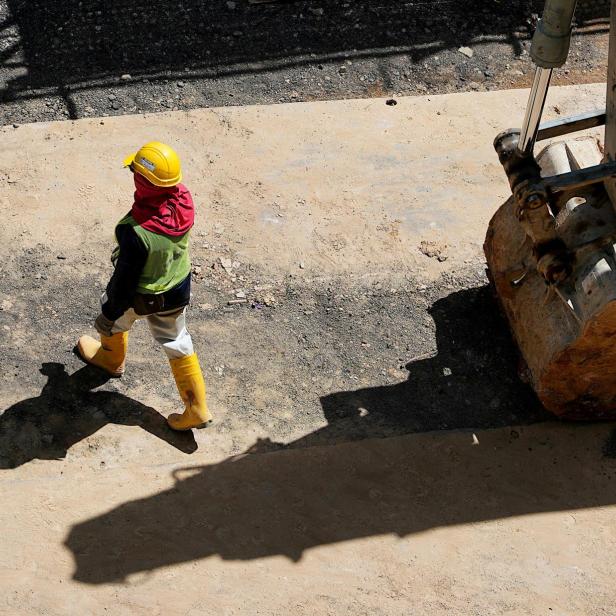 Ein Bauarbeiter mit gelbem Schutzhelm, gelben Gummistiefeln, reflektierender Weste und rotem Tuch um den Kopf geht auf einer Baustelle neben einer grossen Baggerschaufel über einen staubigen Untergrund.