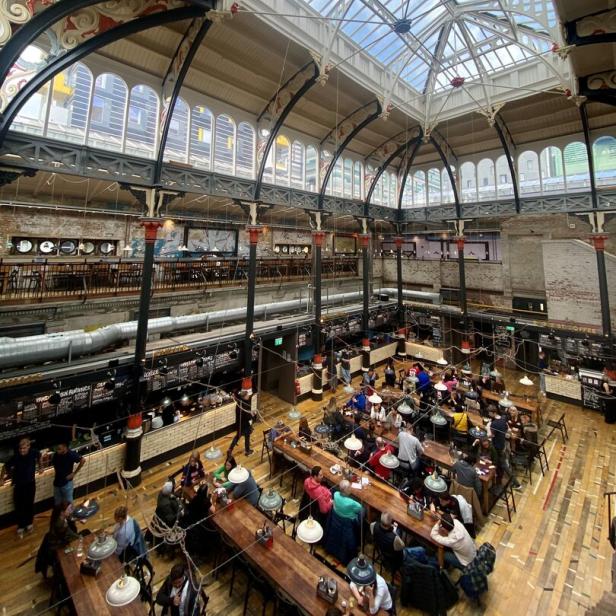 Blick von oben auf eine belebte Markthalle mit vielen Menschen an langen Holztischen.
