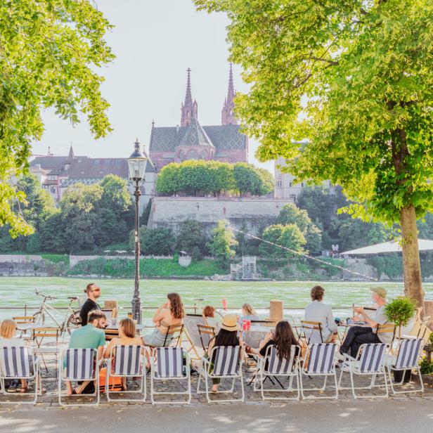 Menschen sitzen in einem Café am Rheinufer in Basel mit Blick auf das Basler Münster.
