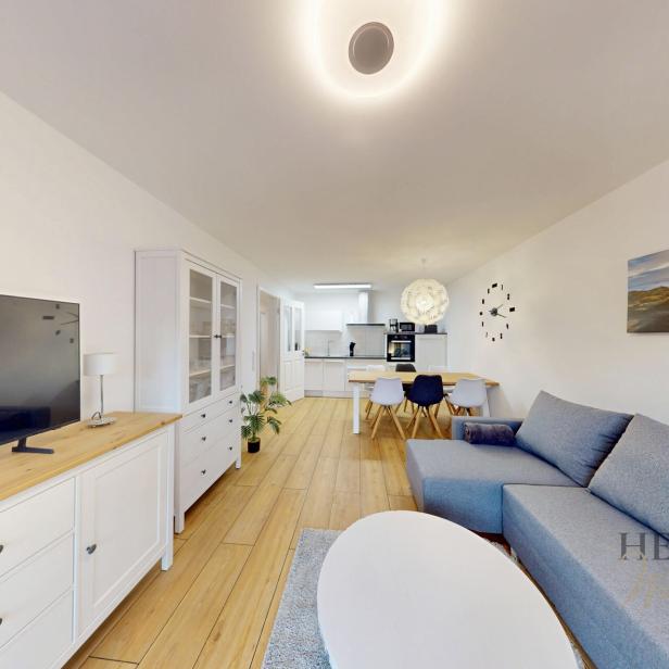 Modern eingerichtetes Wohnzimmer mit offener Küche; im Vordergrund eine graue Ecksofa-Garnitur und ein weisser Couchtisch auf einem hellen Teppich, links ein weisses Sideboard mit Flachbildfernseher und Vitrine, im Hintergrund ein Esstisch mit gemischten Stühlen und eine weisse Einbauküche; an der Wand ein grosses Meeresbild und eine minimalistische Wanduhr; Holzfussboden und helle, freundliche Beleuchtung.