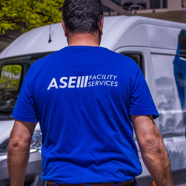 Man sieht einen Mitarbeiter der ASE Facility Services mit gebrandetem T-Shirt von hinten, der auf einen weißen und ebenfalls mit Logo bedrucktem Firmenwagen zugeht.