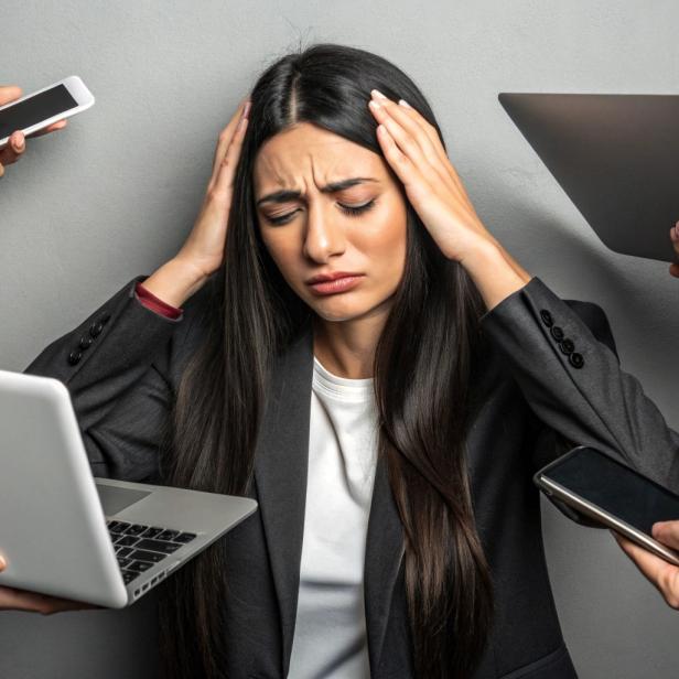 Eine Frau im Businessanzug mit verzweifeltem Gesichtsausdruck greift sich auf den Kopf und ist umgeben von mehreren Laptops.