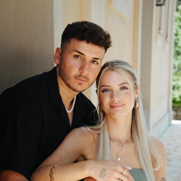 Braunhaariger Mann mit schwarzem Shirt und blonde Frau im grünen Kleid sitzen auf dem Boden und lächeln in die Kamera