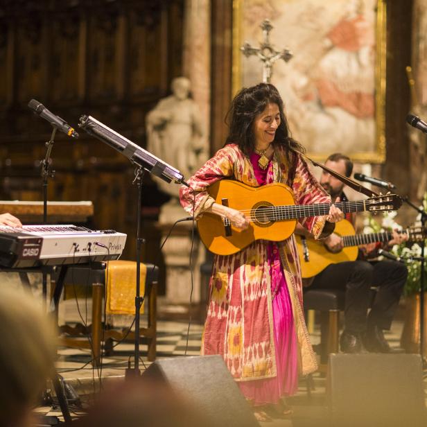Eine Frau steht in bunten Klamotten mit einer Gitarre auf einer Kirchenbühne. 