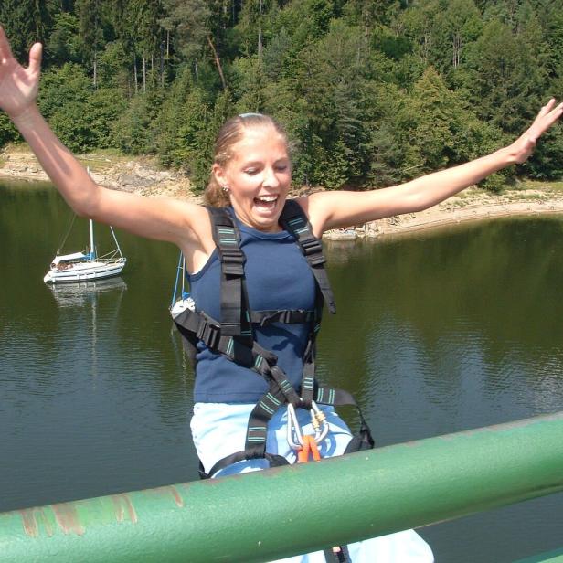 Eine aufgeregte Frau steht mit Bungeegurt auf einer Brücke über einem See.