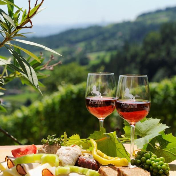 Zwei Gläser Schilcherwein und eine Jause stehen vor einer grünen Hügellandschaft.