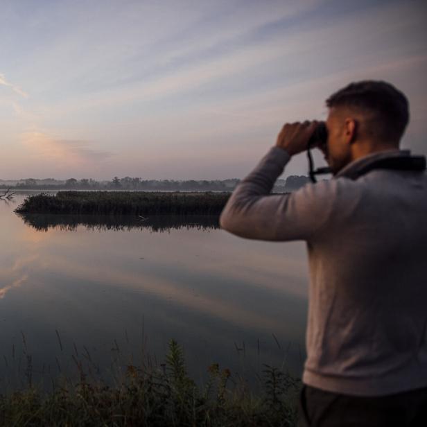 Ein Mann beobachtet mit einem Fernglas die Landschaft am Ufer eines Sees.