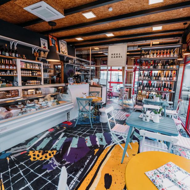 Innenansicht eines Feinkostladens mit Tischen und Regalen voller Produkte.