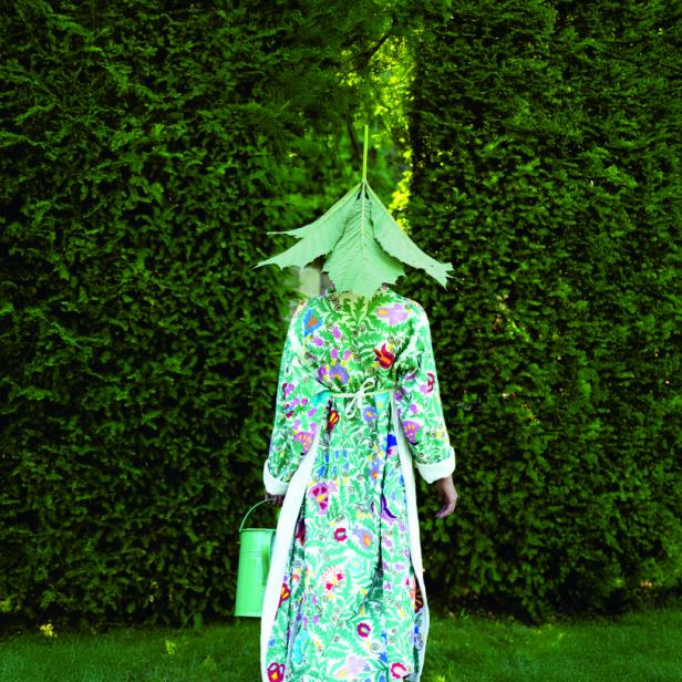 Eine Person in einem floralen Mantel und mit einem Blatt auf dem Kopf steht im Garten.
