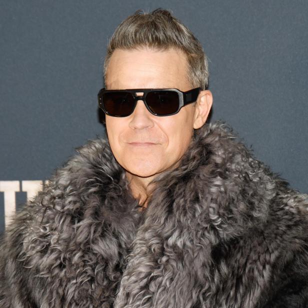 Robbie Williams trägt eine Sonnenbrille und einen grauen Pelzmantel vor einem dunklen Hintergrund.
