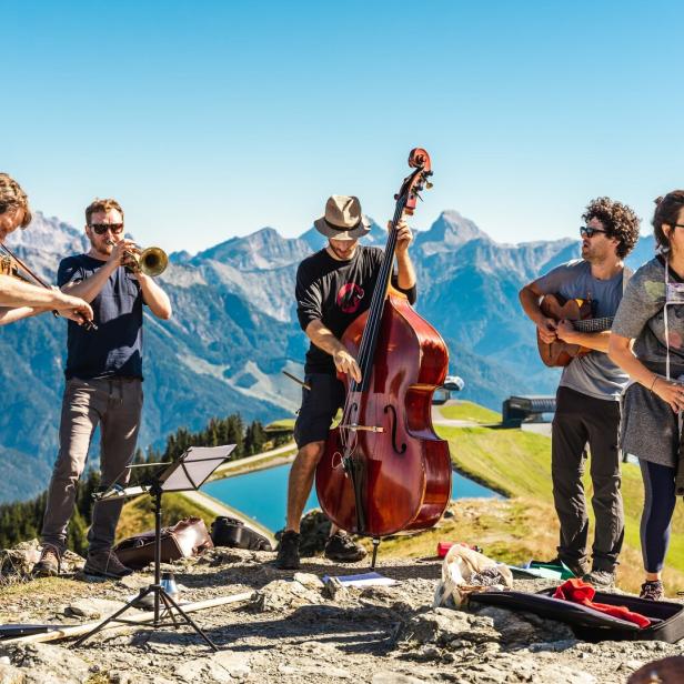 Eine Band spielt im Freien vor einer Bergkulisse mit Geige, Trompete, Kontrabass und Gitarre.