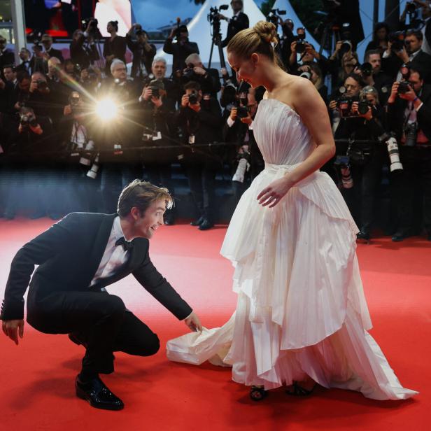 Robert Pattinson hilft einer Frau im weißen Kleid auf dem roten Teppich in Cannes.