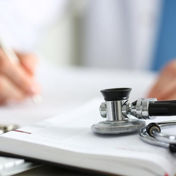 Ein Stethoskop liegt auf einem geöffneten Notizbuch, während eine Person im Hintergrund schreibt.
